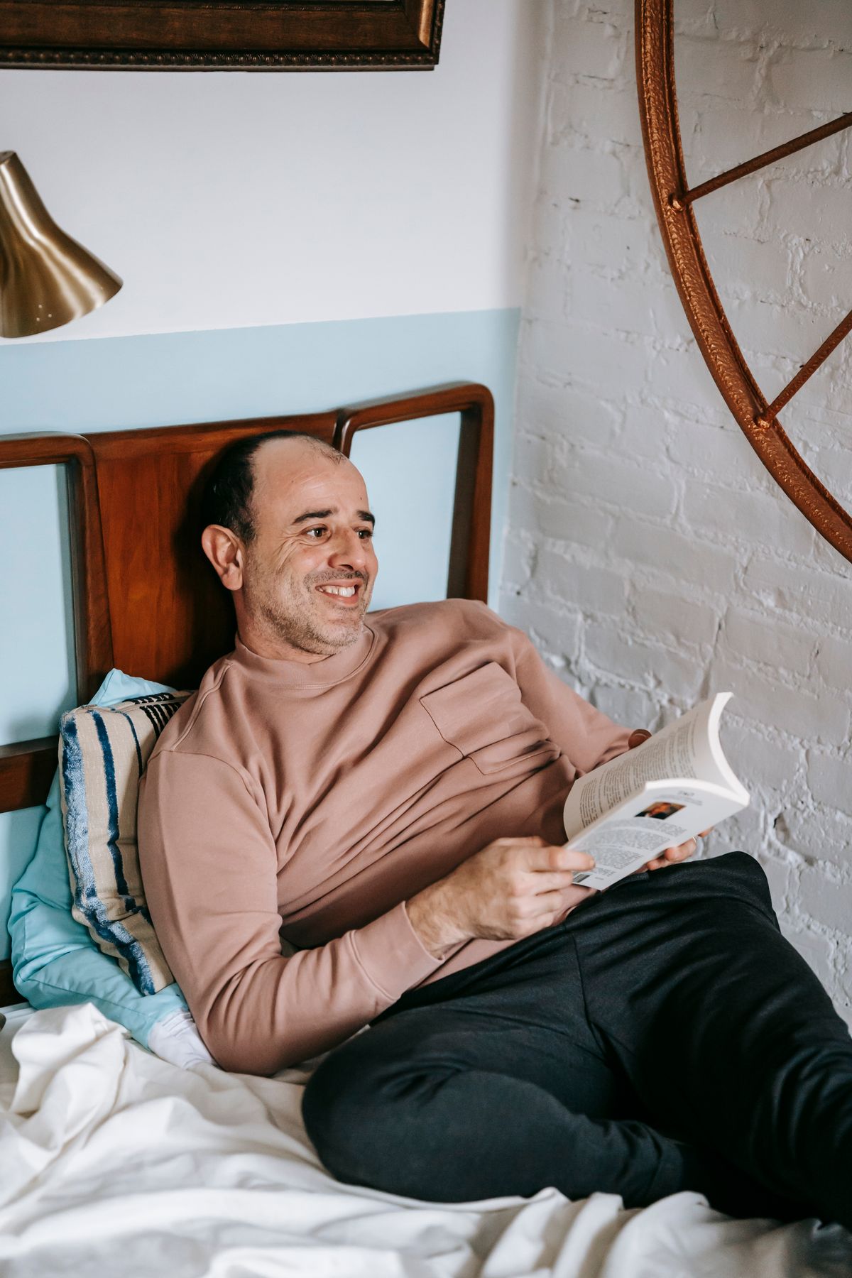 Happy middle aged ethnic male in casual clothes smiling while relaxing on bed and reading interesting book