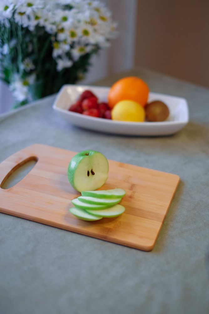 Raw green apple slices and a halved apple on a wooden cutting board; fruit tray behind.