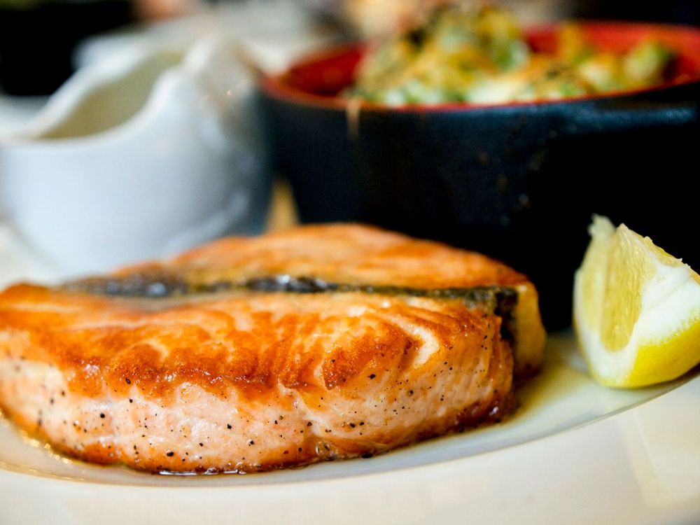 A browned salmon fillet on a white plate, with side food in the background.