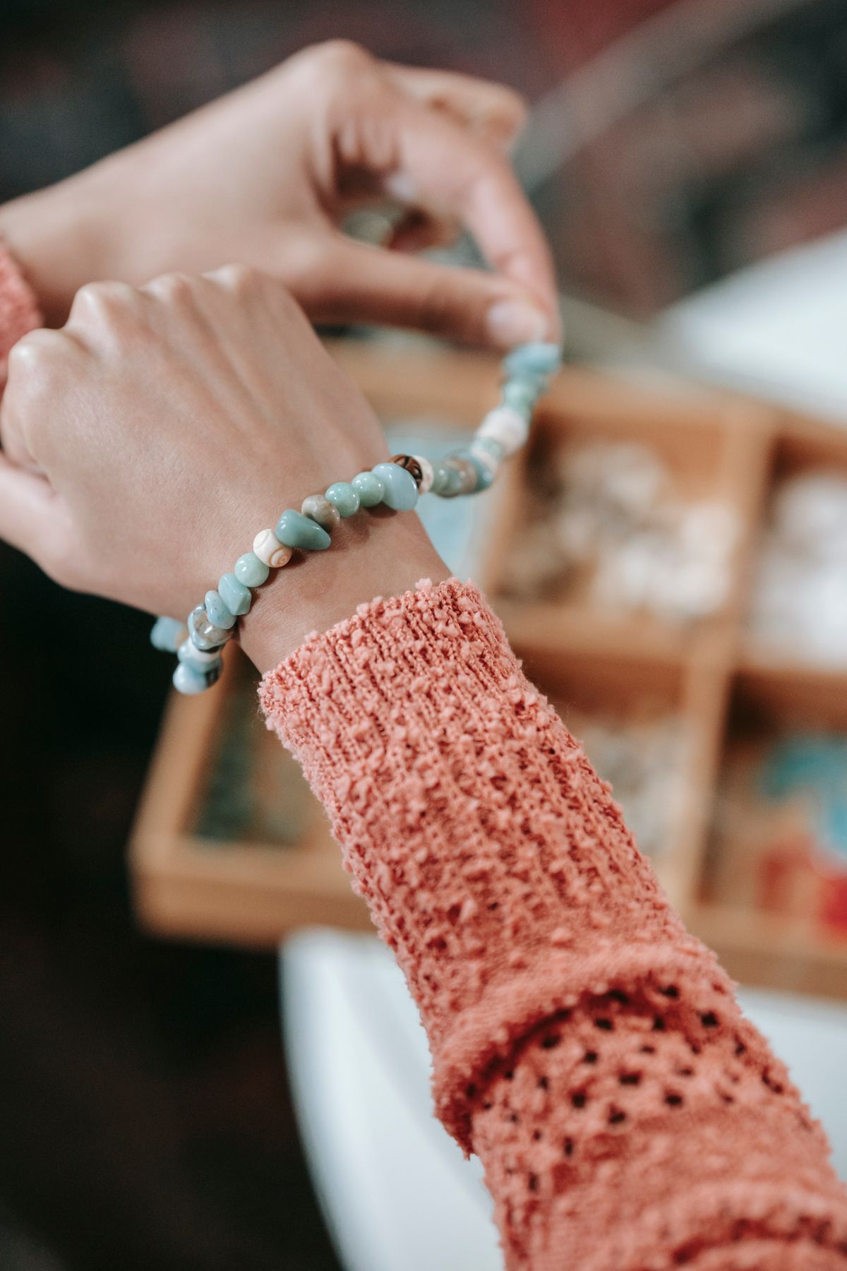 From above of crop anonymous female with handmade bracelet on hand sitting at table with container full of supply on blurred background