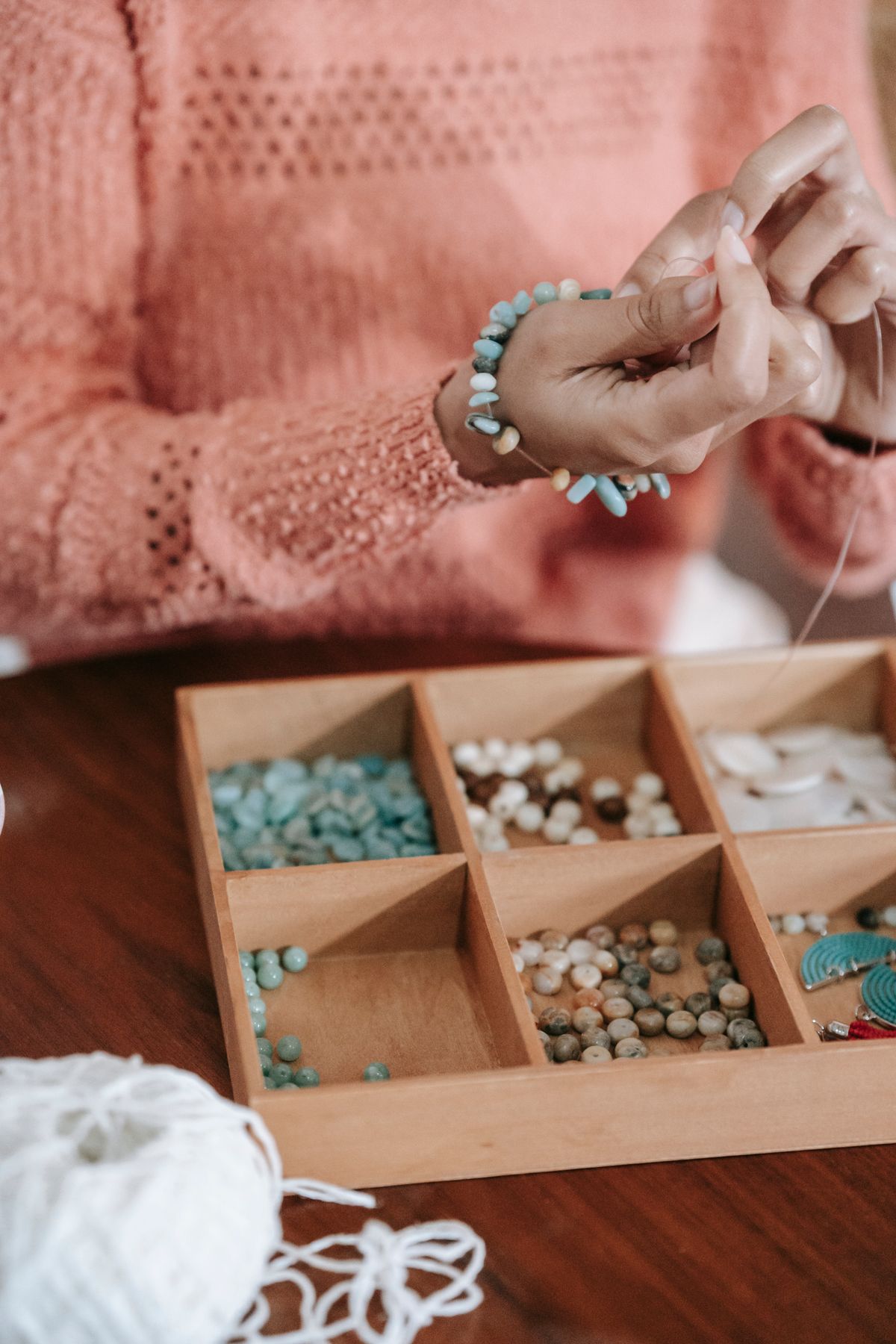 Crop anonymous ethnic female master sitting at table with box with various beads and making bracelet