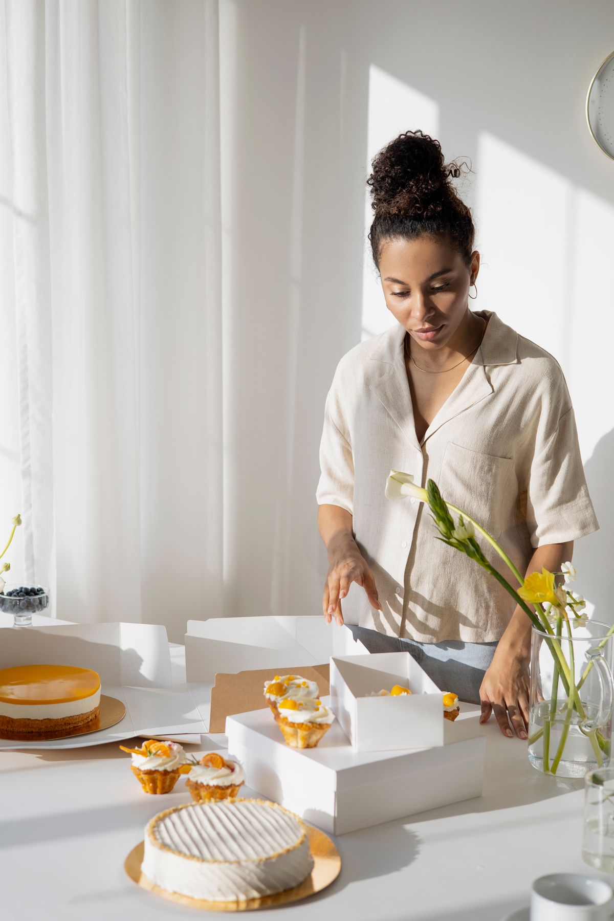 A female pastry chef carefully organizes cakes and pastries in a sunlit kitchen space.