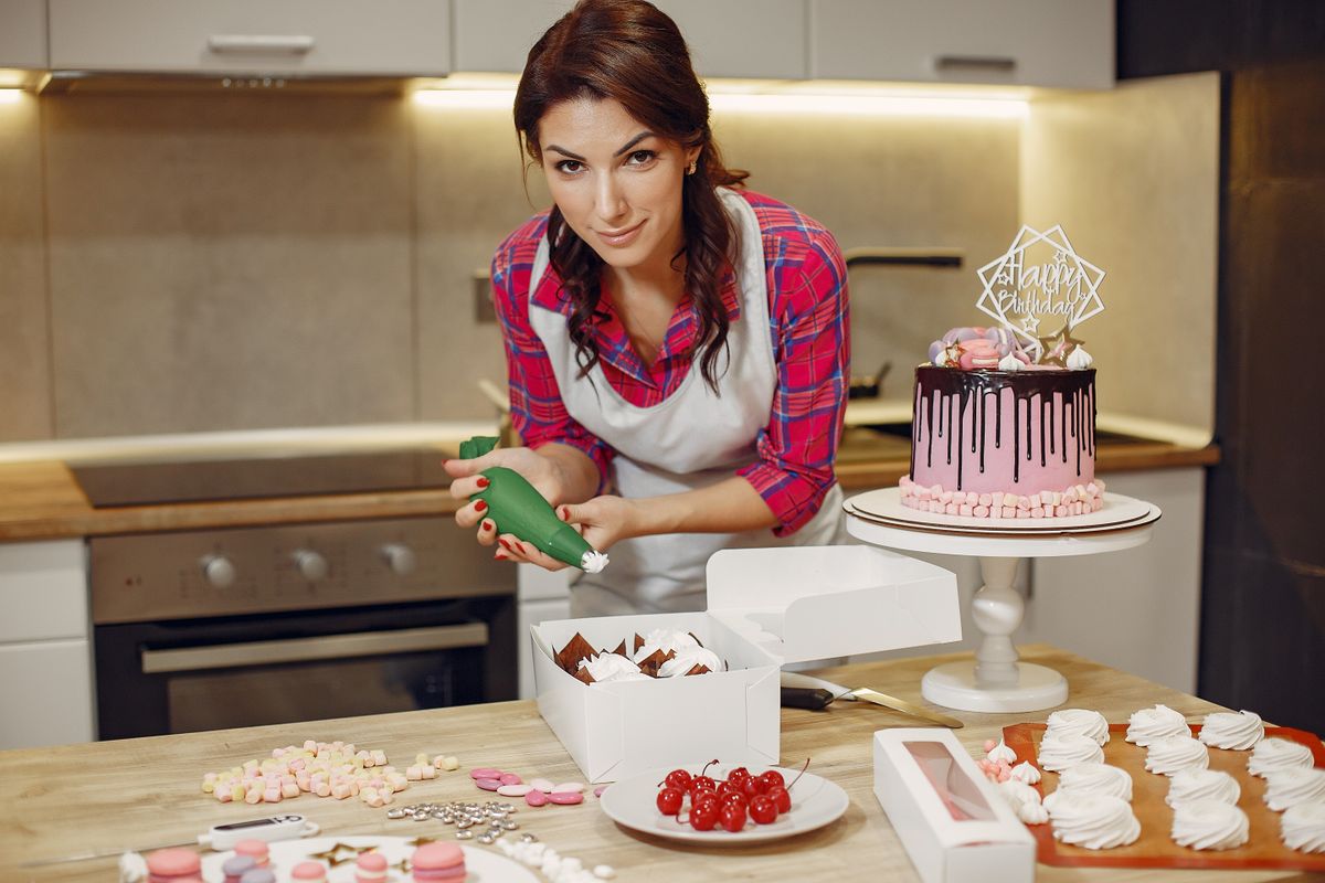 A woman in an apron decorates a cake with icing in a contemporary kitchen setting.