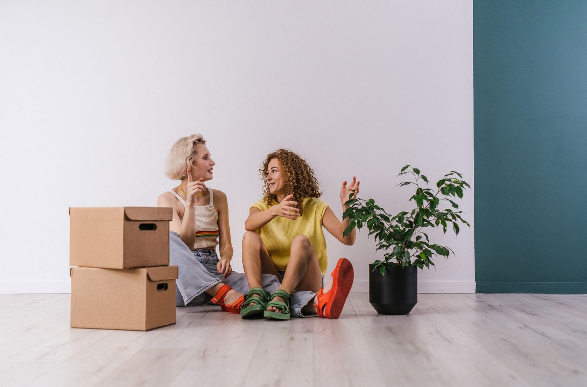 Two women sitting on the floor, unpacking boxes while talking in a new apartment.
