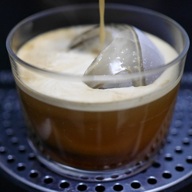 A coffee stream entering a short clear glass containing ice cubes, on a machine drip tray.