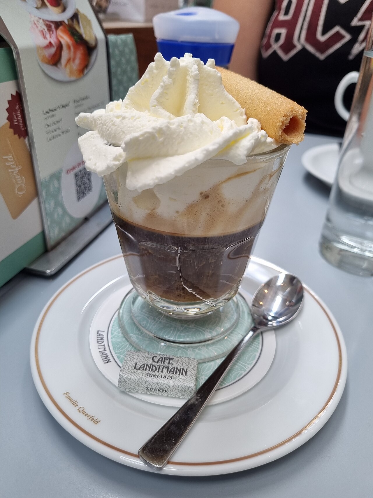 Dark coffee in a clear footed glass under whipped cream and a rolled wafer, with a spoon on its saucer.