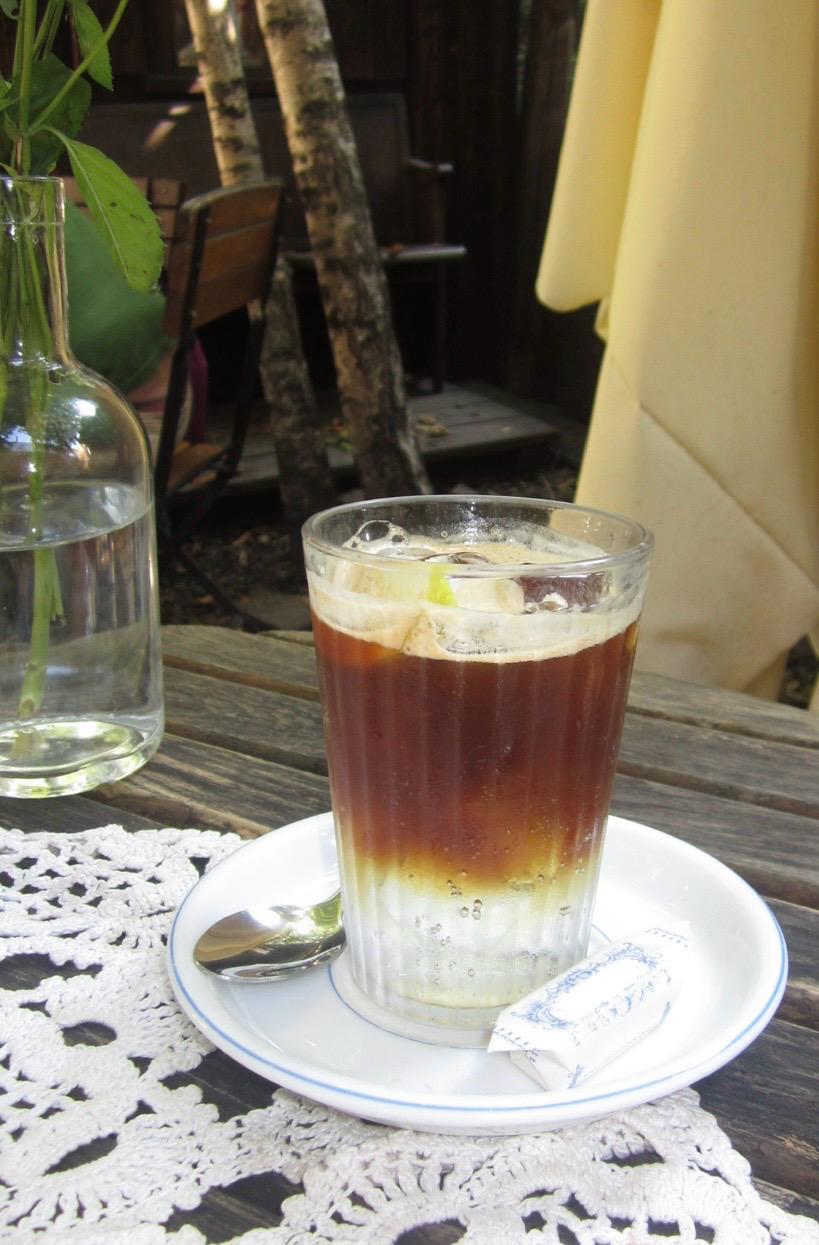 Tall clear glass of layered espresso tonic with ice and a small garnish, on a white saucer.