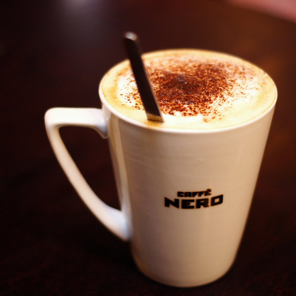 White mug holding a pale foamy white-chocolate mocha with brown dusting and a spoon.