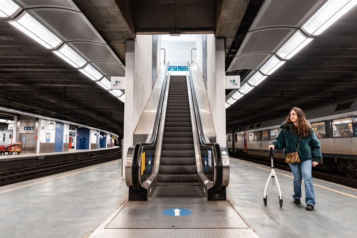 A woman with a rollator at an empty modern train station platform near an escalator.