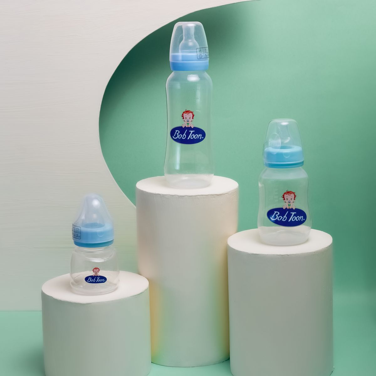 Three baby bottles displayed on cylindrical pedestals with a green backdrop, highlighting modern design.