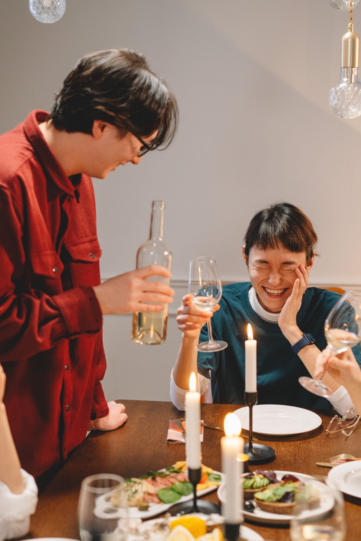 Two friends laughing over dinner with wine and candles, enjoying a cozy night indoors.
