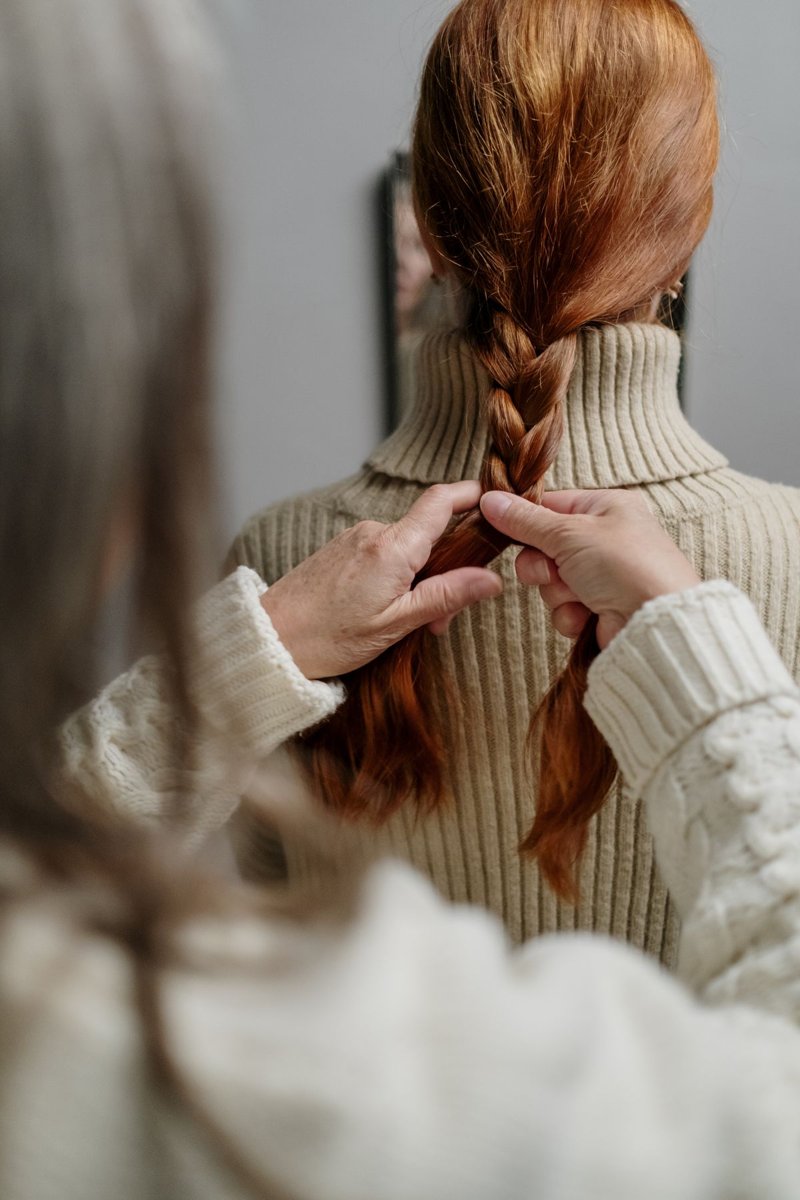 Hands braiding a red-haired woman’s hair from behind.
