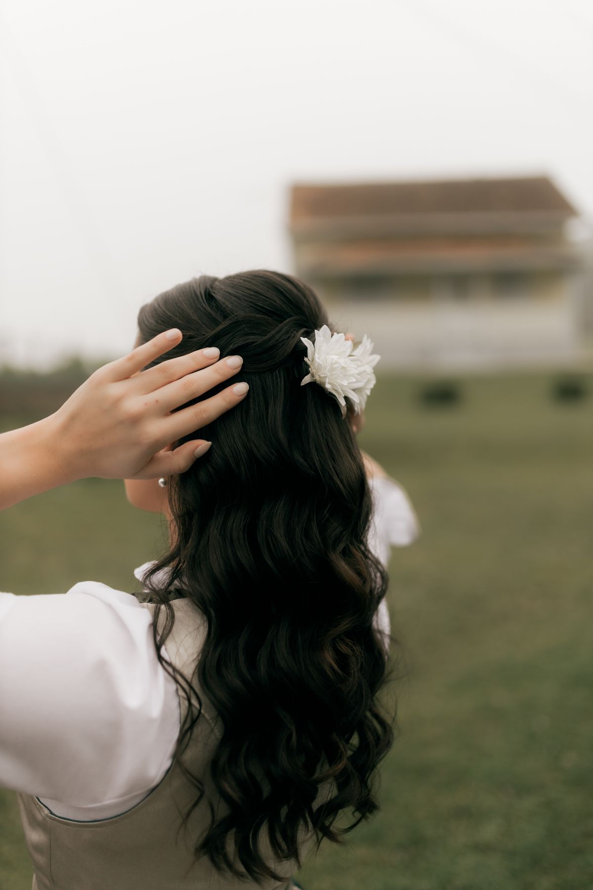 Woman with long dark hair and flower accessory outdoors, gently touching her hair.