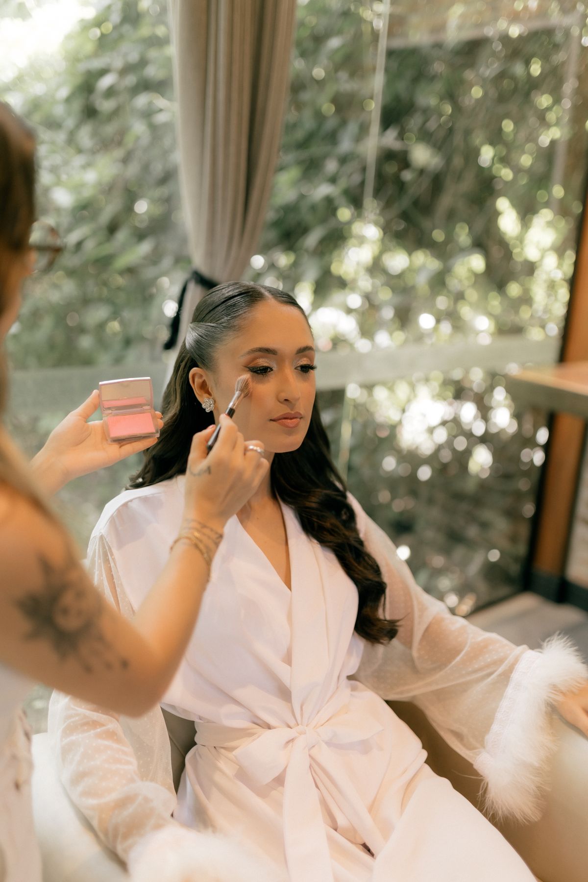 A bride in a white robe getting makeup applied by an artist, surrounded by natural light.