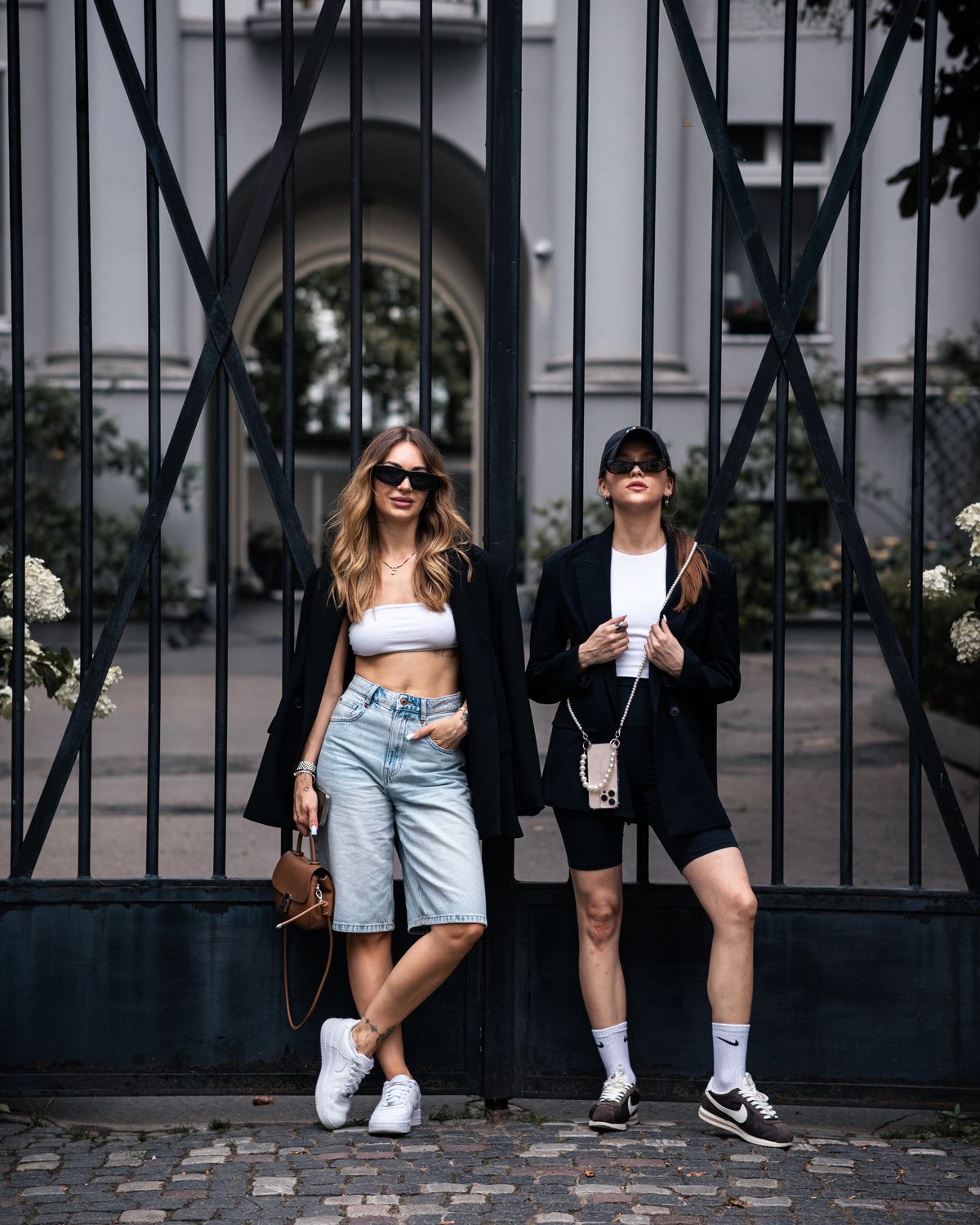 Two stylish women posing in urban fashion wear against a decorative gate.