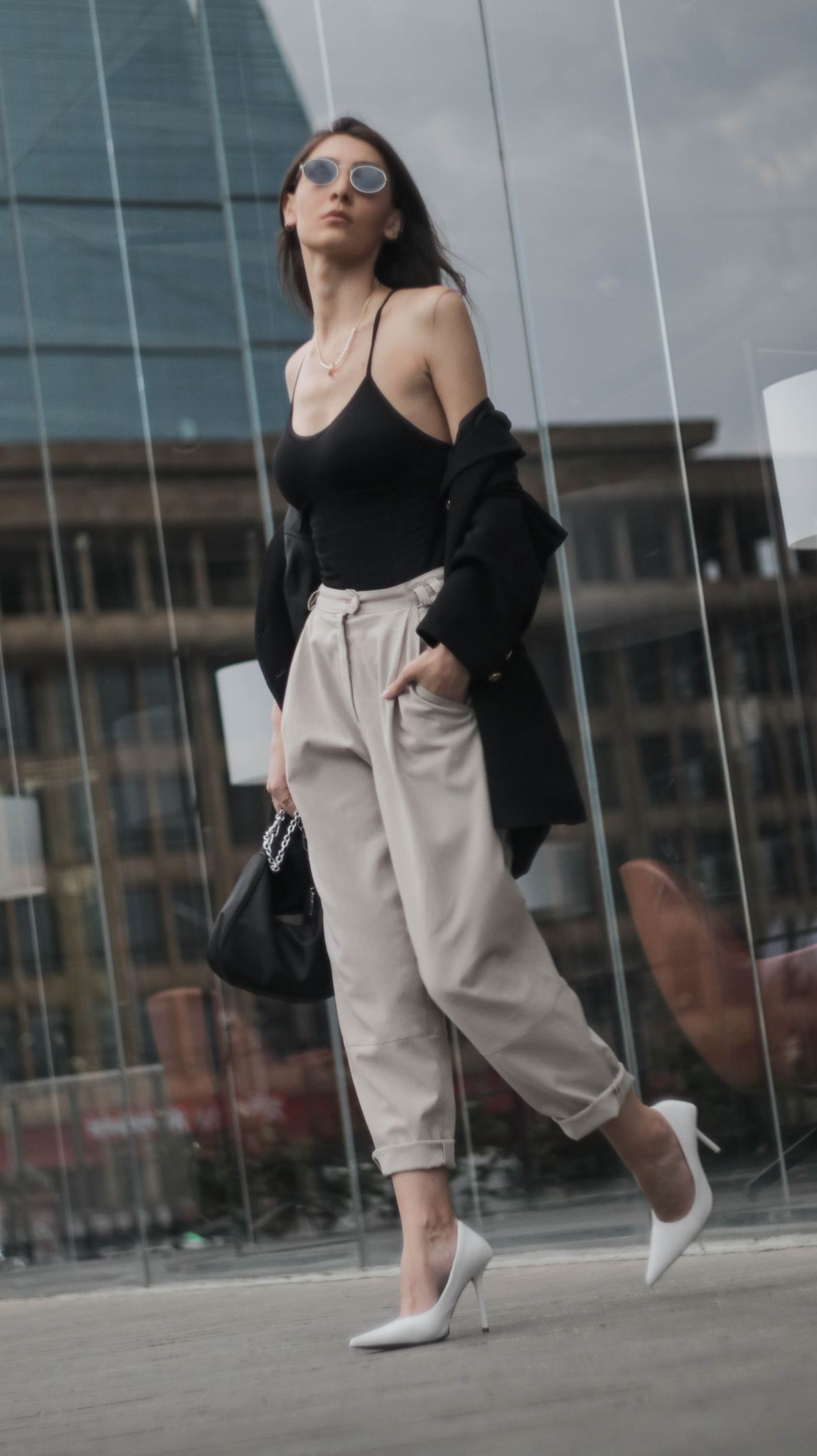 From below of confident young female in stylish clothes and sunglasses strolling on street near modern glass building at daytime