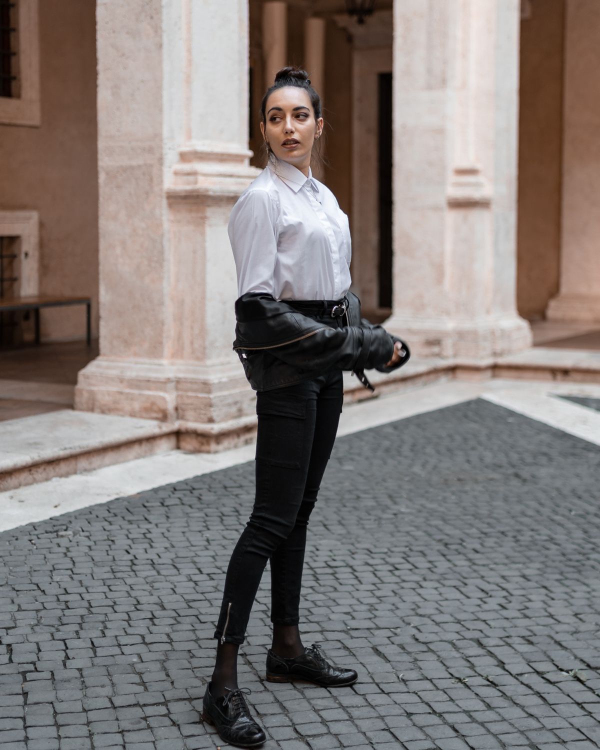 Full body of young female in trendy outfit standing near stone building with columns and looking away