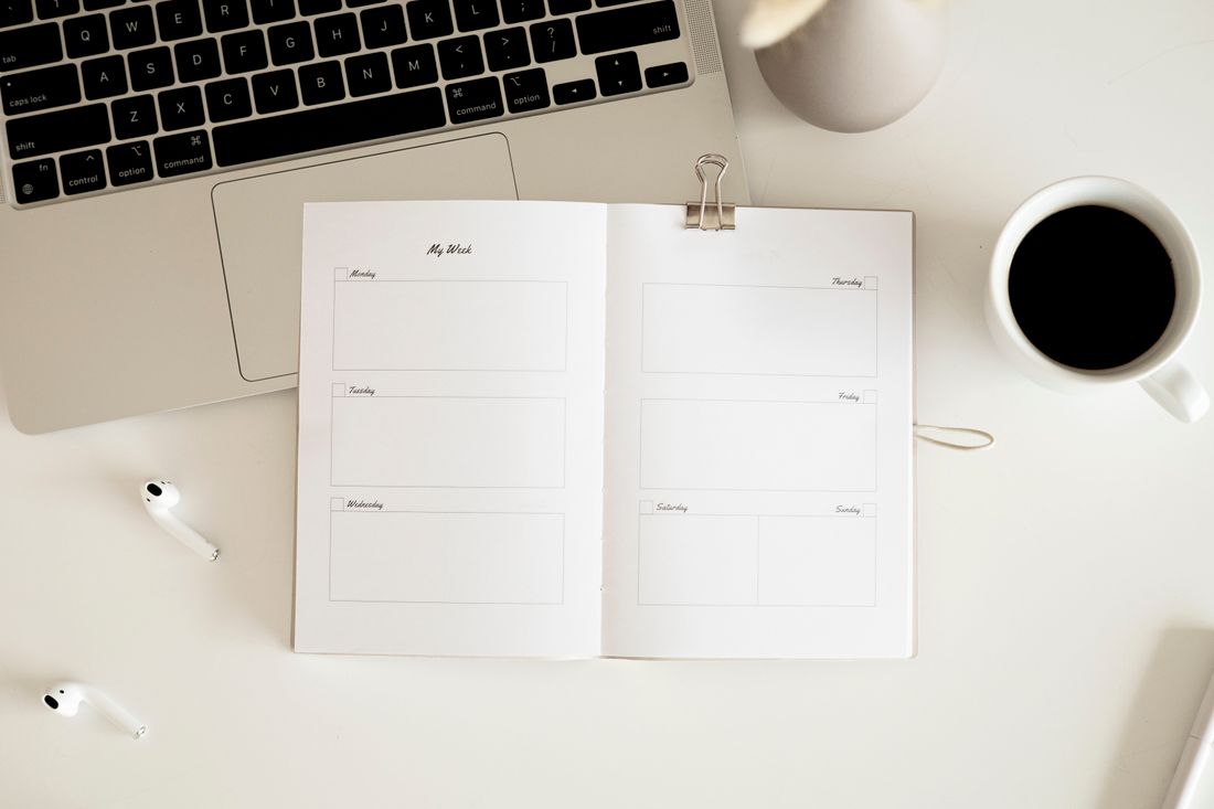 An open planner beside coffee, a laptop and earbuds.