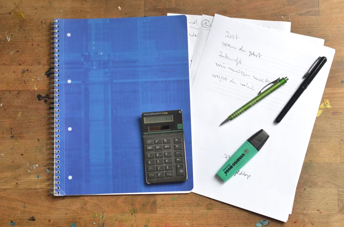 A notebook, calculator, markers and pens on a wooden desk.