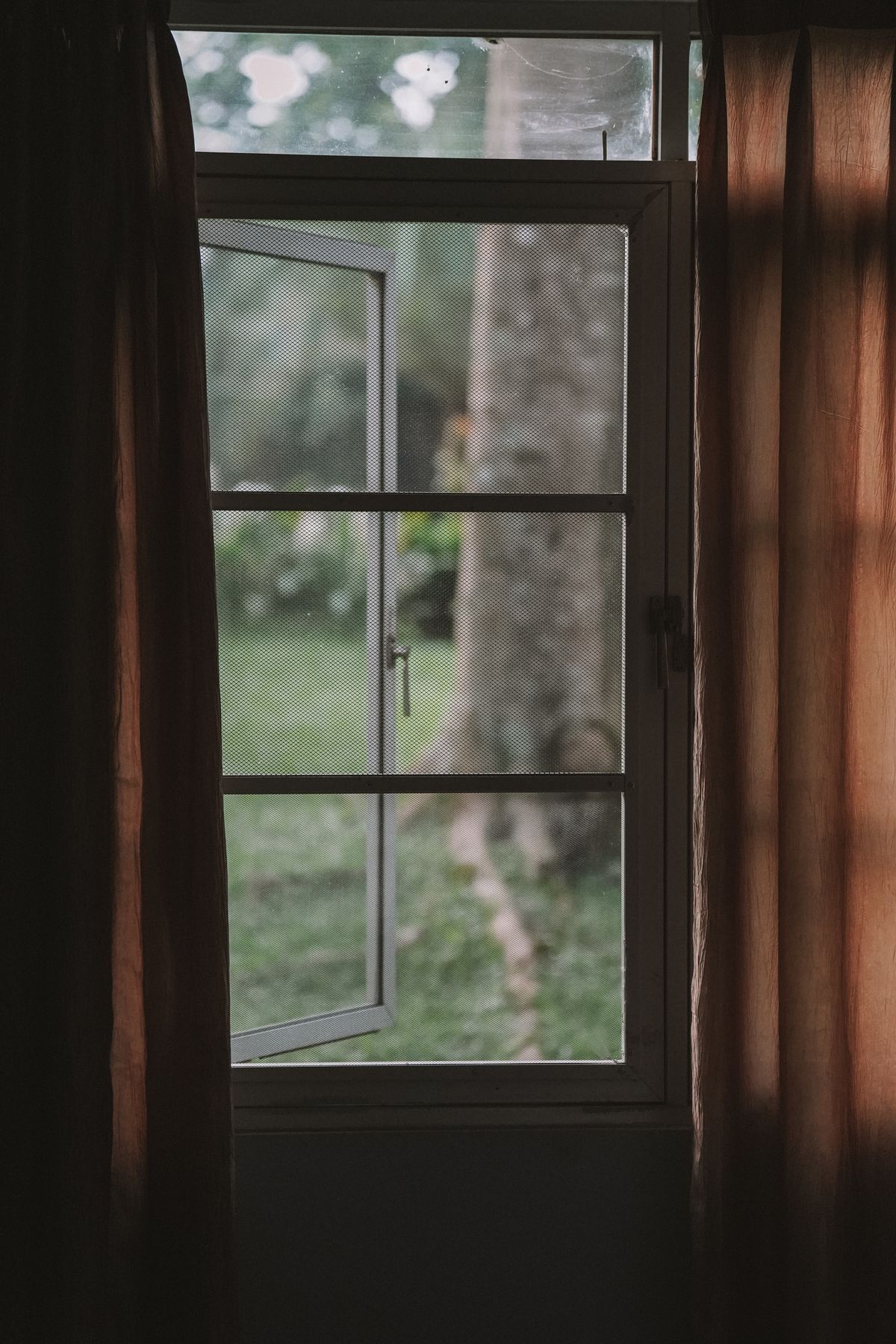 A serene view through an open window with brown curtains revealing a lush garden in West Java, Indonesia.